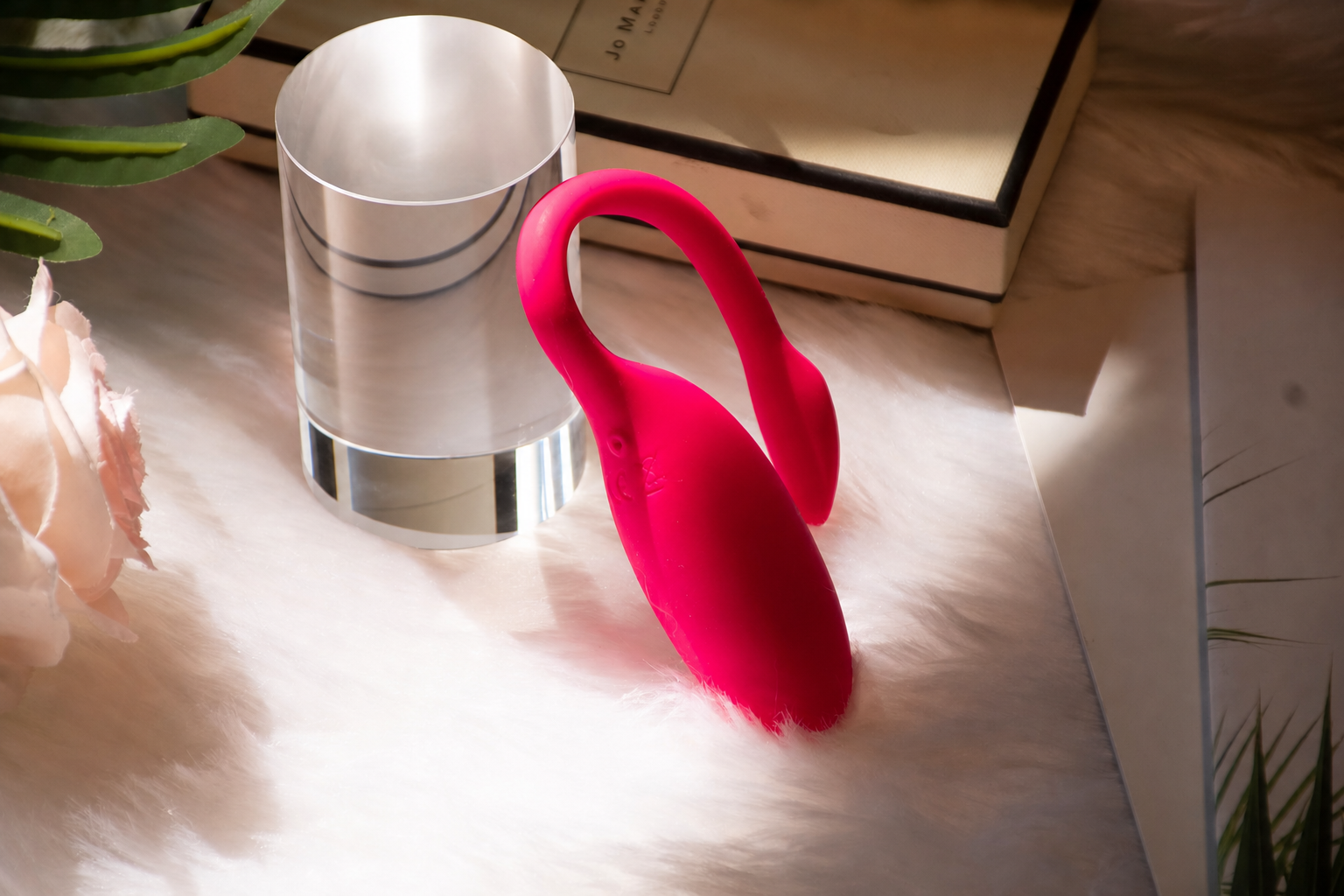 Magic Flamingo pink wearable vibrator beside a polished clear cylinder on a soft bedside surface