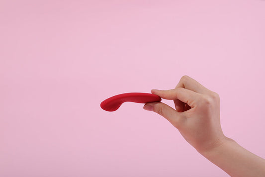 Hand holding a red curved vibrator against a pink background.