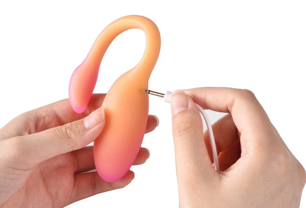 Hands connecting a white pin-style charging cable to an orange wearable vibrator