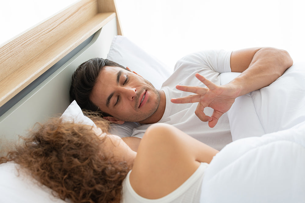 Couple lying in bed while one person makes an OK gesture.