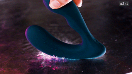 Hand holding a dark green prostate massager above colorful powder.