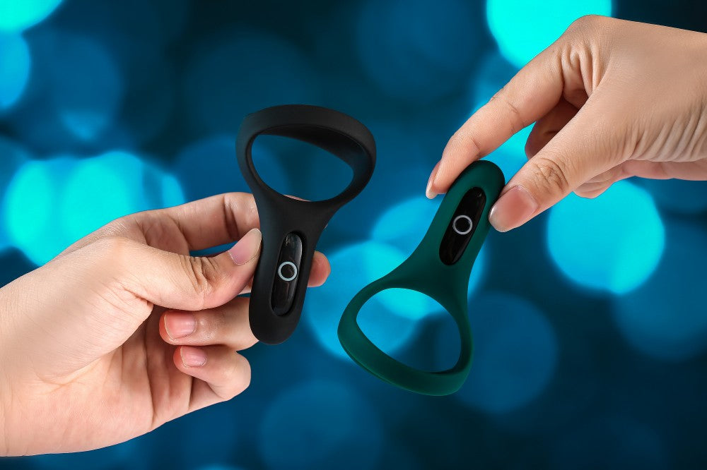 Hands holding two black and green cock rings against a teal background.