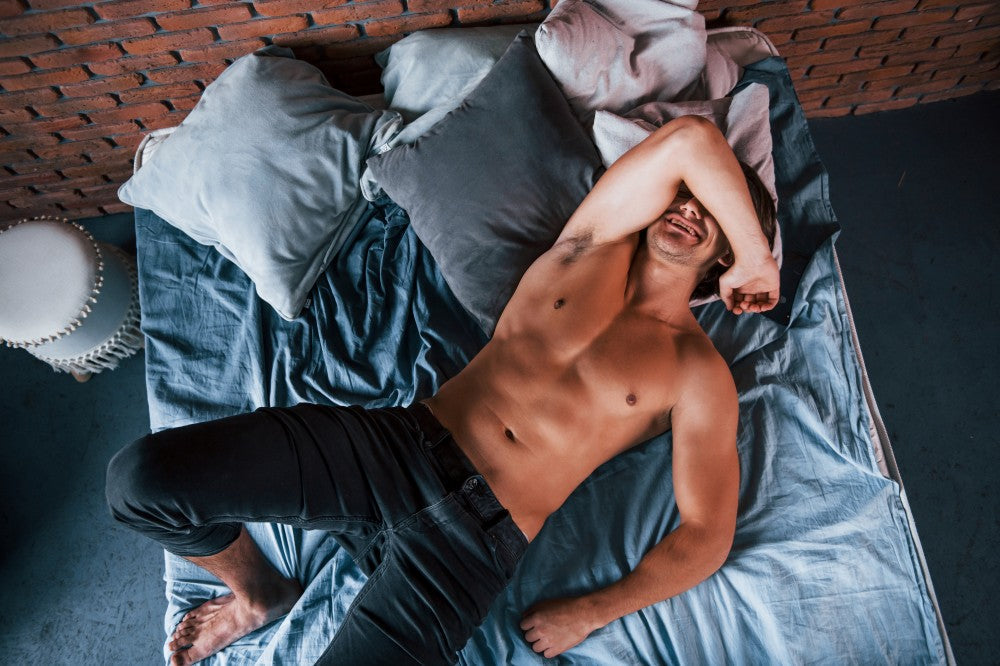 Shirtless man lying on a bed with one arm over his face.
