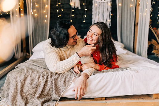 Smiling couple sitting together on a bed under string lights.